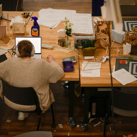 High-angle shot of a student working on a laptop at a wooden table with drafting tools and papers.