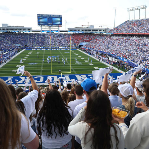 Fans fill a packed stadium as players line up on the field at a University of Kentucky football game, with the crowd waving white rally towels.