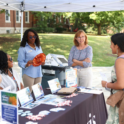 People speak with visitors at an outdoor information table under a tent on campus, with brochures, handouts and a landfill waste display.