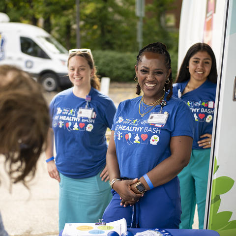Three people wearing blue “Be Healthy Bash” shirts stand beside a mobile health unit and greet an attendee at an outdoor event. A table with event materials is in the foreground, and a branded vehicle is visible in the background.