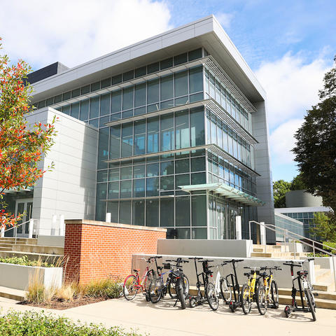 A modern, multi-story glass academic building sits on a landscaped campus, with a small tree showing autumn colors in the foreground and a bike rack filled with bicycles near the entrance on a sunny day.