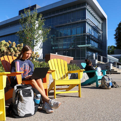 Student sitting in a bright yellow Adirondack chair outside a modern glass campus building, working on a laptop with a backpack and water bottle nearby, while other students sit and study along the walkway on a sunny day.