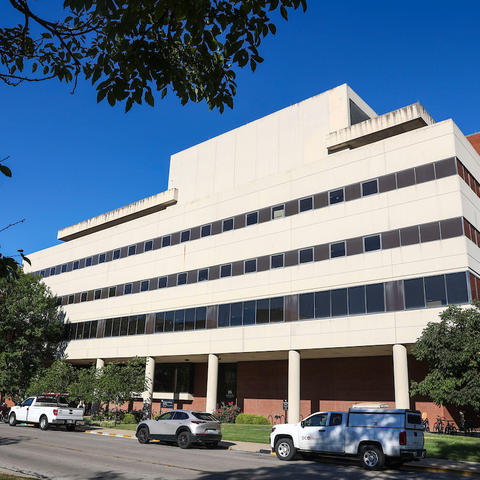 This is an exterior photo of the College of Nursing Building at UK, a multistory building with a white stone front lined with columns along a street with parked vehicles and a clear blue sky. 