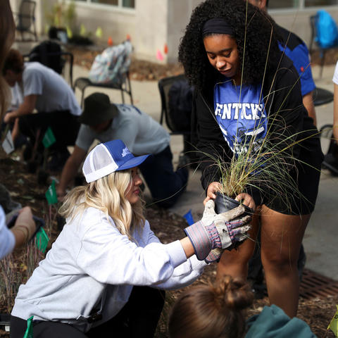 A Black woman standing and a white woman crouching, preparing to put a plant into a garden.