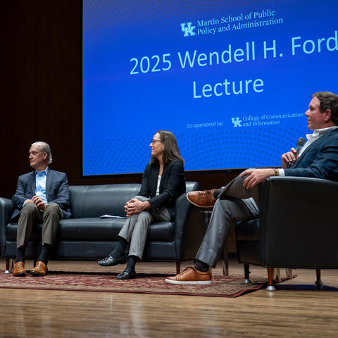 Four panelists sit on a stage during a public lecture and discussion at the University of Kentucky. One speaker gestures while talking from an armchair, while the other panelists listen from a sofa and adjacent chairs. A large screen behind them.