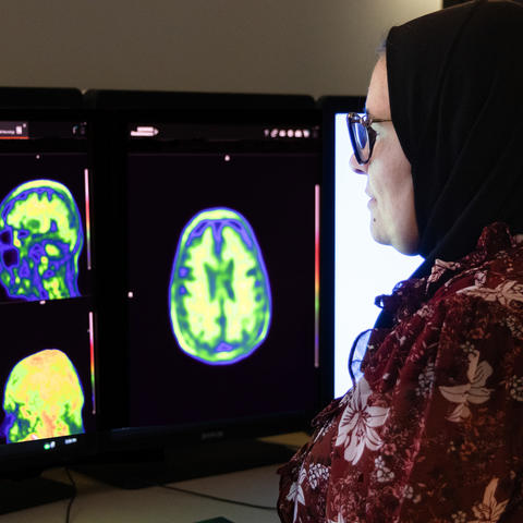 A woman wearing glasses and a headscarf studies colorful brain scan images displayed on two computer monitors in a medical research setting.