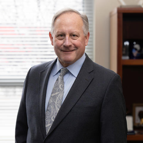A middle-aged man in a suit and patterned tie stands smiling in an office, with window blinds behind him and a bookshelf with awards and objects to his side.