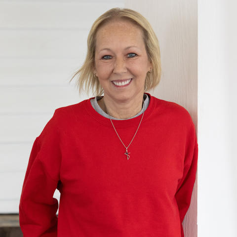Vikki Lane smiles at the camera while standing in front of a white wall. She is wearing a red sweater with a gray collar and a silver necklace.
