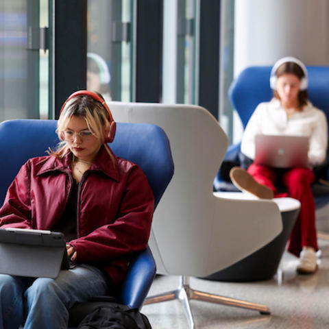 Two students wearing headphones sit in chairs by large windows, one using a tablet in the foreground while another works on a laptop in the background.