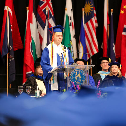 A student in a blue graduation cap and gown speaks at a podium on a stage, with faculty seated behind and numerous international flags displayed in the background.