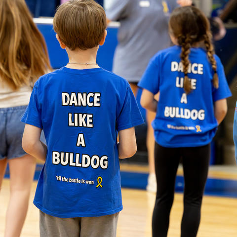 Children wearing matching blue T-shirts that read “Dance Like a Bulldog” stand in a gymnasium, facing away from the camera as they participate in a group activity.