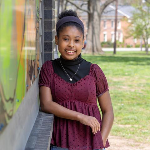 A teenage girl stands outdoors, leaning against a colorful mural wall, with grass, trees, and campus buildings in the background.