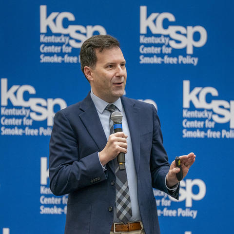 Person holding a microphone and speaking in front of a blue backdrop reading “KCSP Kentucky Center for Smoke-free Policy.”