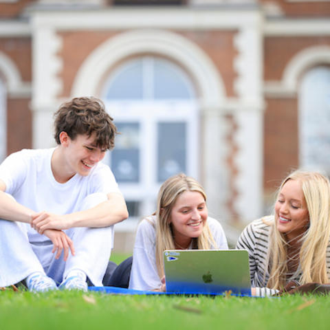 Three students sitting on grass outside a campus building, smiling and looking at a laptop together.