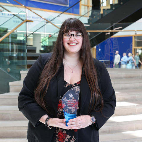 A person with long dark hair, wearing a black blazer over a floral dress, holds a glass award while standing on indoor steps in a modern building with blue accents.