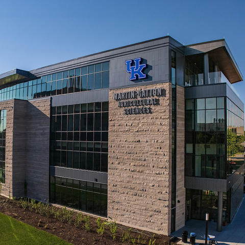 Aerial view of the University of Kentucky’s Martin-Gatton Agricultural Sciences building, featuring a stone-and-glass exterior and a large UK logo under a clear blue sky.