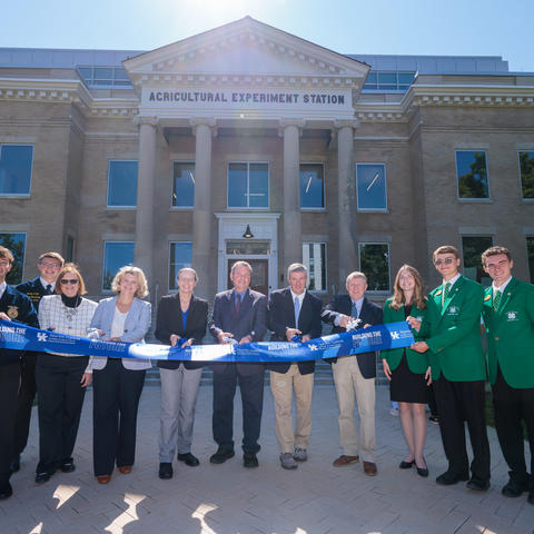 A group of people stands in front of the University of Kentucky Agricultural Experiment Station building during a ribbon-cutting ceremony. The historic brick building features a columned entrance and a sign reading “AGRICULTURAL EXPERIMENT STATION." 