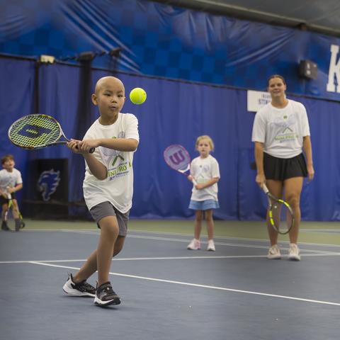 A child swings a tennis racquet to hit a bright yellow tennis ball on an indoor court at the University of Kentucky. Three other young tennis players and an instructor stand in the background holding racquets. Blue court curtains and large “KENTUCKY” lett