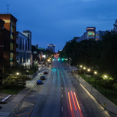 Elevated view of a campus street at dusk, with illuminated buildings and trees lining the road as a vehicle’s red taillights streak through the foreground.