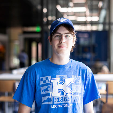 A person wearing a University of Kentucky cap and blue K Week t-shirt stands in a campus study area with tables and chairs in the background.