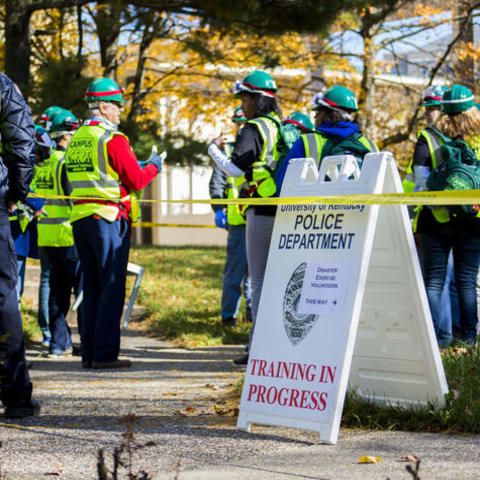 Emergency responders standing outside by yellow tape next to a sign that says "UK Police Department, Training in Progress"