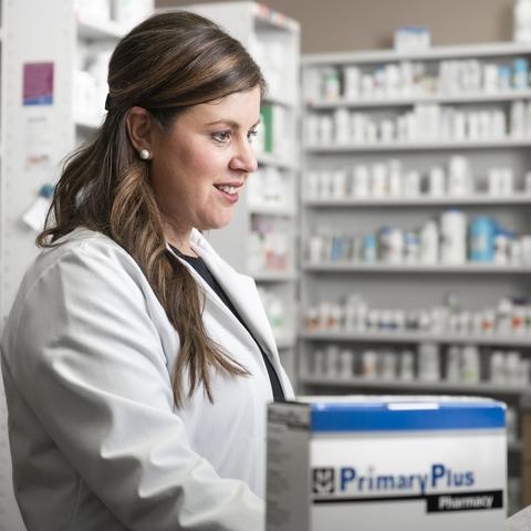 Pharmacist in a white lab coat standing behind a pharmacy counter with shelves of medication in the background.