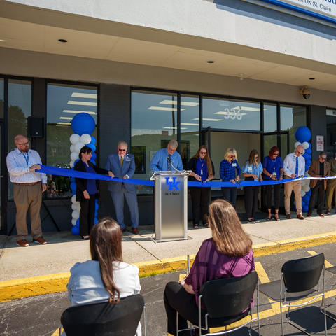 A ribbon-cutting ceremony takes place outside a row of storefronts at a shopping center on a sunny day. A long blue ribbon stretches across the sidewalk as a large group of people — many wearing matching blue shirts — stand in a line behind it.