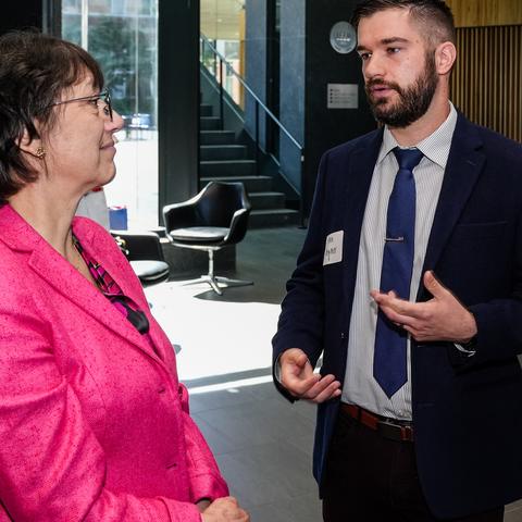 A young white man in a dark blue suit and navy tie presents a research poster to NIH director Monica Bertagnolli, a middle-aged white woman wearing a pink suit.