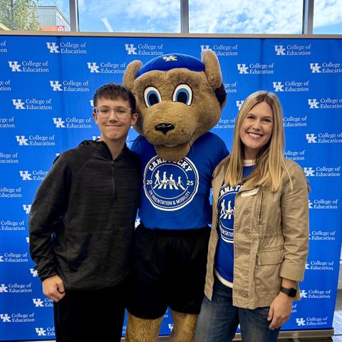 University of Kentucky backdrop with two people posing beside a costumed mascot wearing a blue shirt and cap featuring a circular logo.