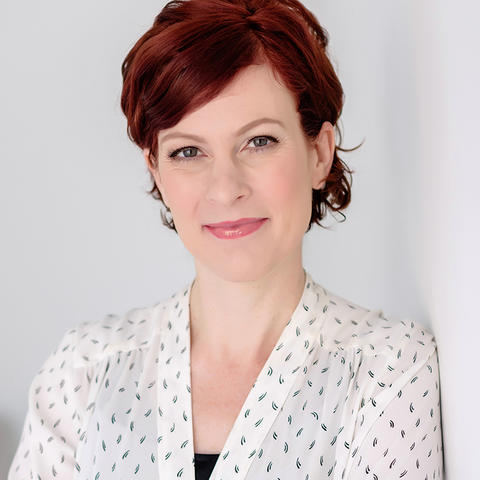 A person with short, wavy red hair stands against a plain light-colored wall, wearing a white blouse with a subtle dotted pattern.