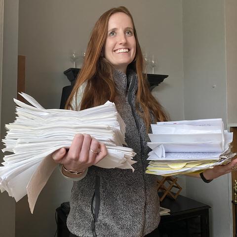 A person standing indoors holds large stacks of white papers and envelopes in both arms, with documents fanned out and slightly overflowing, suggesting a heavy workload or paperwork overload.
