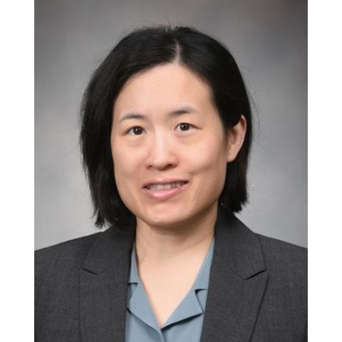 Professional headshot of a person with short dark hair wearing a gray blazer and light blue collared shirt, posed against a gray studio background.