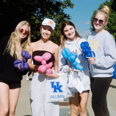 Four people stand outdoors holding colorful balloon creations, including flowers and an animal-shaped balloon.