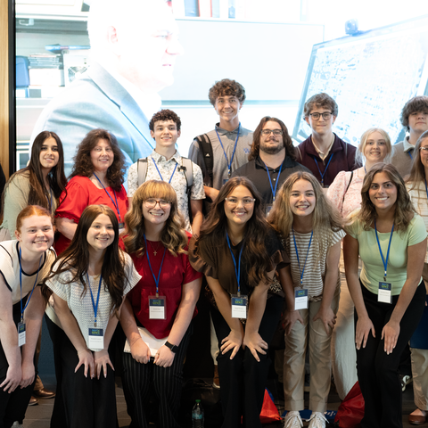 A group of students wearing conference lanyards pose for a photo in front of a large display screen.