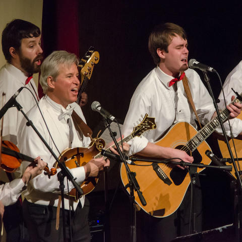 A musical group of four men performing on stage, each wearing white shirts and bow ties; two are playing acoustic guitars, one is playing a violin, one is playing a mandolin, and all are singing into microphones under warm stage lighting.