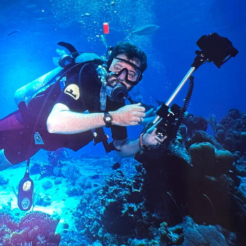 A scuba diver swims above a coral reef in clear blue water while holding an underwater camera rig. 