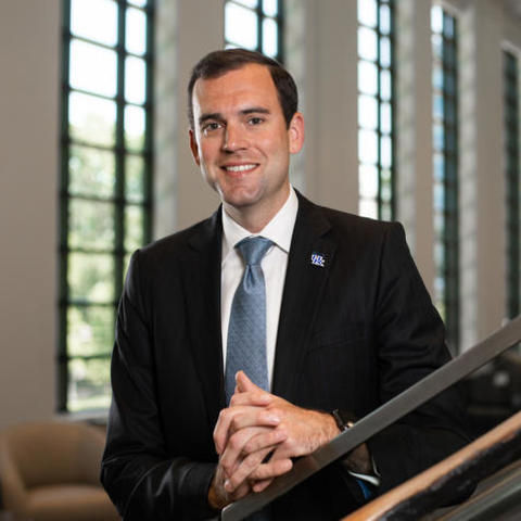Portrait of an individual in business attire standing indoors near a staircase, hands clasped. The person wears a dark suit, light blue tie, and UK lapel pin. Large windows in the background let in natural light, with seating visible along the wall.