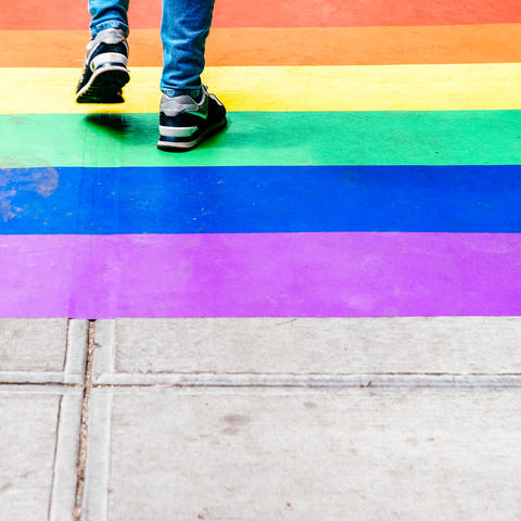 The image shows the lower legs of a teenager walking over a rainbow sidewalk; they're wearing jeans and black sneakers.