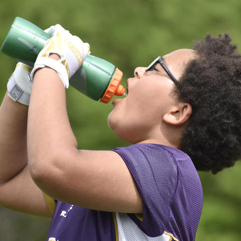 Student athlete wearing a football practice jersey and gloves drinks water from a sports bottle outdoors during warm weather.