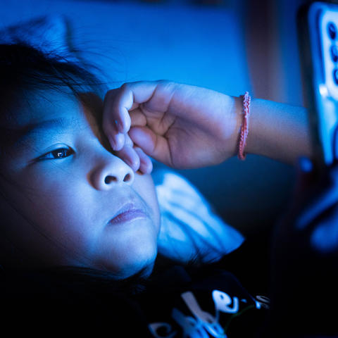 A child lies in bed in a dark room, holding a smartphone close to their face. Blue light from the phone illuminates the child's face and hand while the rest of the room remains dimly lit.