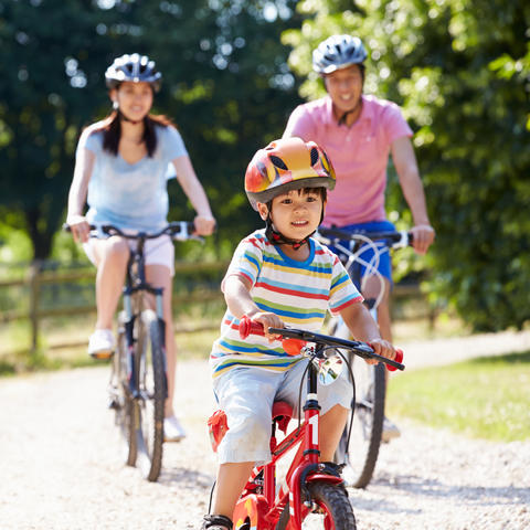 A child wearing a helmet rides a red bicycle along a sunlit gravel path, with two helmeted adults riding bikes behind them in a wooded park setting.