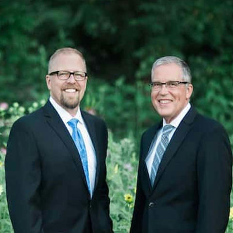 Photo of two white men wearing glasses and dark suits in front of a garden background