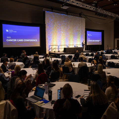Attendees at round tables facing a stage where speakers present at the 2025 Cancer Care Conference hosted by UK Markey Cancer Center, with illuminated backdrop and multiple display screens.
