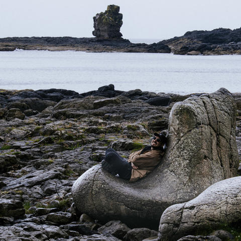 A person sits on a large rock formation along a rugged shoreline during a visit to a coastal landscape.