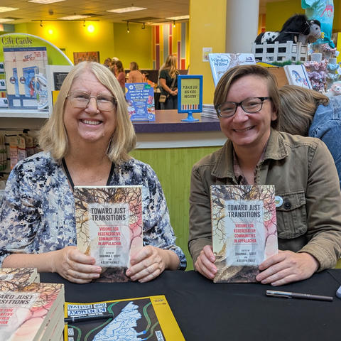 Two people sit at a table at an indoor event, smiling while holding up copies of a book. A display of books and posters is visible behind them.