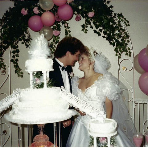 Photo of a young white couple at their wedding. A multi-tiered cake is in the foreground; the couple stand behind it, facing each other with foreheads touching and big smiles on their faces. They're framed by a canopy of foliage and pink balloons. 