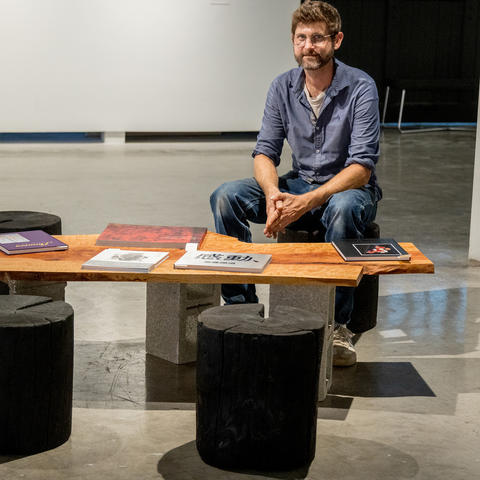 A person sits behind a low wooden bench displaying colorful books and printed materials, with two black stools in front, inside a gallery space.