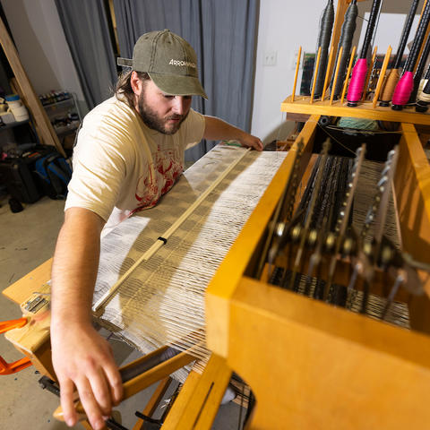 White male in a white t-shirt wearing a ball cap, seated at a loom. 