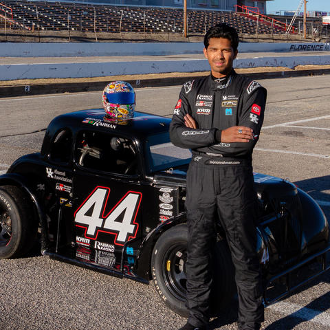 A person in a black racing fire suit stands with arms crossed in front of a black Legend race car marked with the number 44. The car features multiple sponsor logos, including the UK Alumni Association, and a colorful racing helmet rests on the roof.