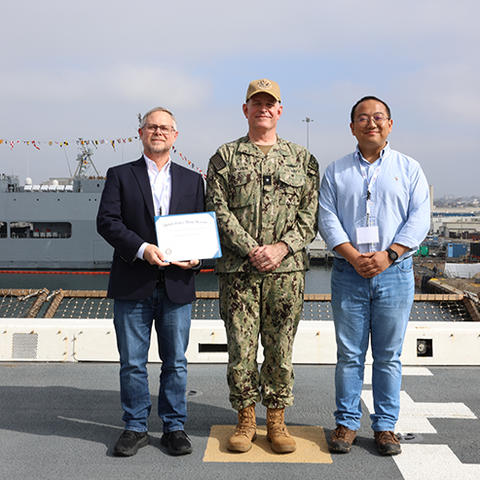 three men, one in a blazer holding a certificate, one in Navy uniform and one in a button-down shirt, stand together on the deck of a ship with another Navy ship behind them
